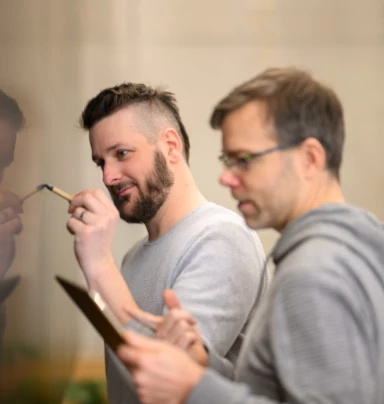 Alex gibt Hannes Informationen von seinem Tablet, während Hannes diese an einer Tafel zusammenfasst.